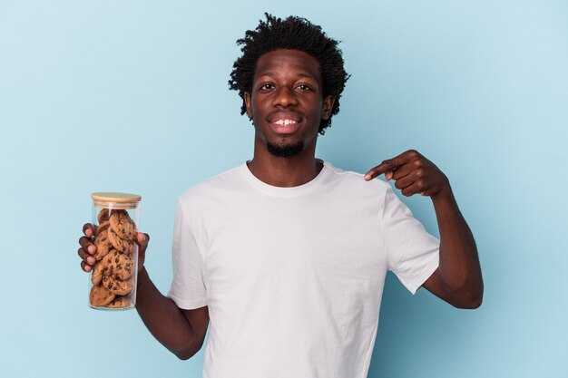 파란색 배경에 격리된 초콜릿 칩 쿠키를 들고 있는 젊은 아프리카계 미국인 남성이 셔츠 복사 공간을 손으로 가리키며 자랑스럽고 자신감이 넘칩니다.