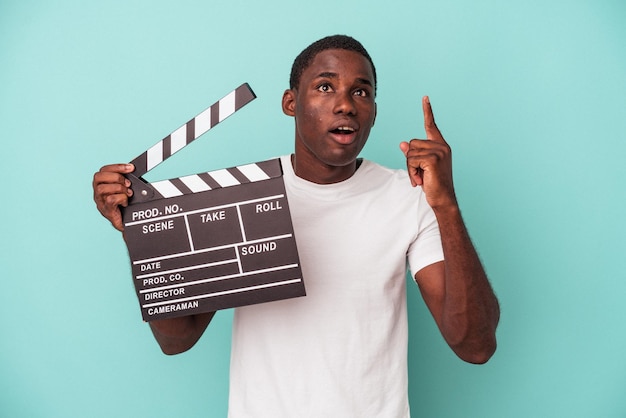 Young African American man holding clapperboard isolated on blue background pointing upside with opened mouth.