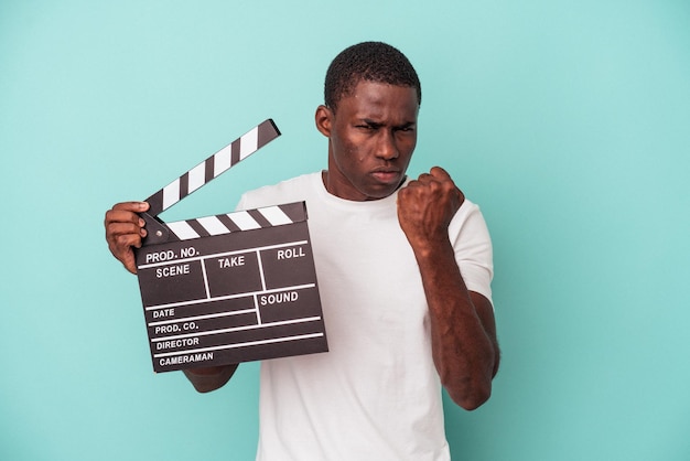 Young African American man holding clapperboard isolated on blue background showing fist to camera, aggressive facial expression.