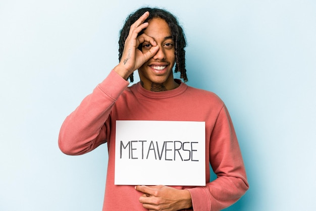 Young African American man holding metaverse placard isolated on blue background excited keeping ok gesture on eye