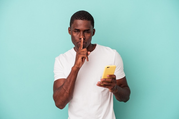 Young African American man holding a mobile phone isolated on blue background keeping a secret or asking for silence