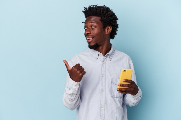 Молодой афро-американский человек, держащий мобильный телефон, изолированный на синем фоне, указывает пальцем далеко, смеясь и беззаботно.