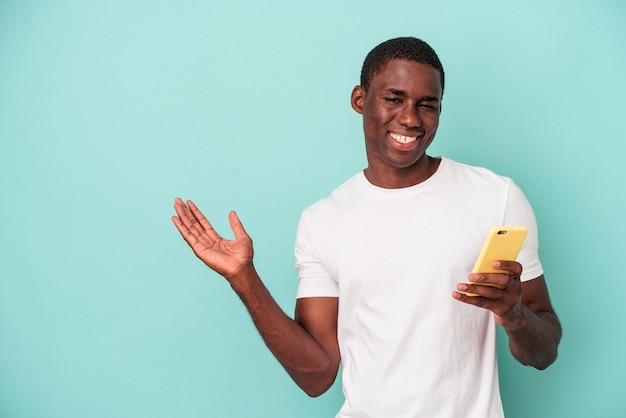 手のひらにコピースペースを示し、腰に別の手を保持している青い背景で隔離の携帯電話を保持している若いアフリカ系アメリカ人の男性。