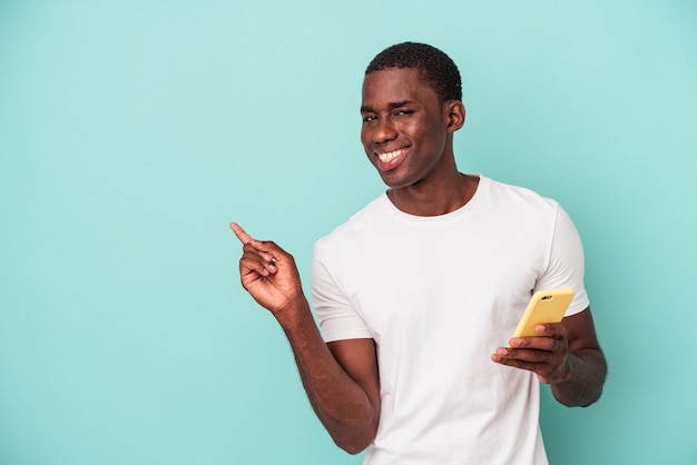 Young African American man holding a mobile phone isolated on blue background smiling and pointing aside, showing something at blank space.