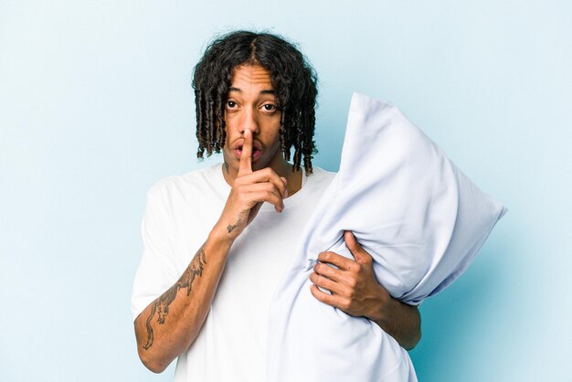 Young African American man holding a pillow isolated on blue background keeping a secret or asking for silence