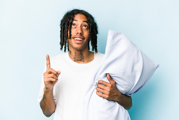 Young African American man holding a pillow isolated on blue background pointing upside with opened mouth