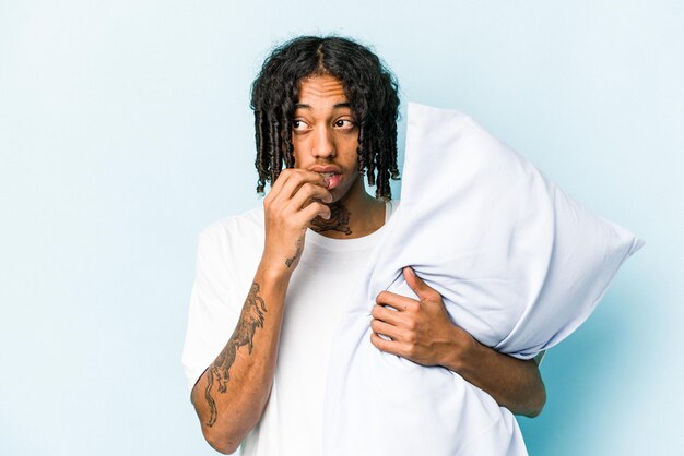 Young African American man holding a pillow isolated on blue background relaxed thinking about something looking at a copy space