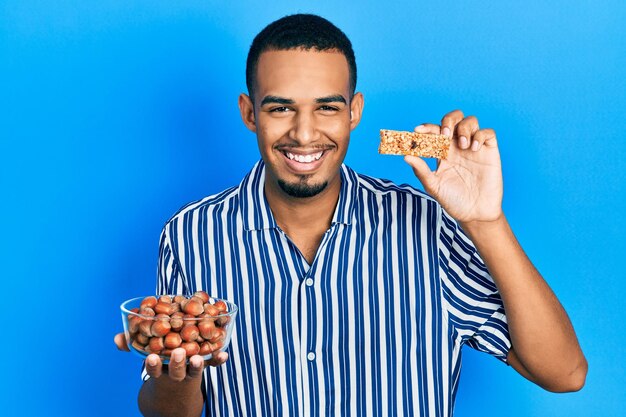 Young african american man holding raw hazelnuts and cereal bar smiling and laughing hard out loud because funny crazy joke