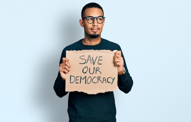 Young african american man holding save our democracy protest banner smiling looking to the side and staring away thinking