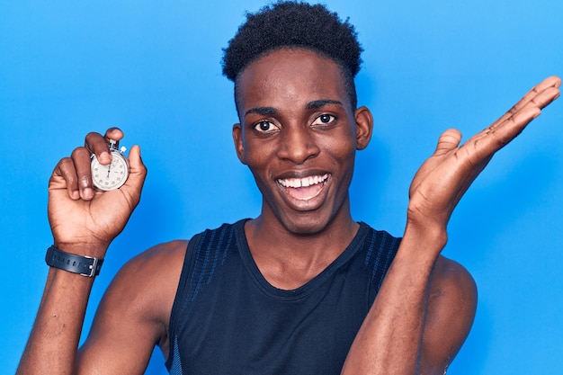 Young african american man holding stopwatch celebrating victory with happy smile and winner expression with raised hands