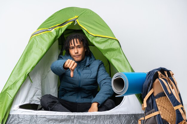 Giovane uomo afroamericano dentro una tenda verde di campeggio che mostra pollice giù con l'espressione negativa