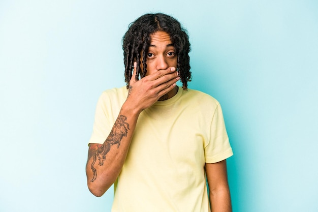 Young African American man isolated on blue background covering mouth with hands looking worried