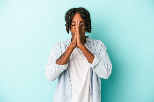 Photo young african american man isolated on blue background holding hands in pray near mouth, feels confident.