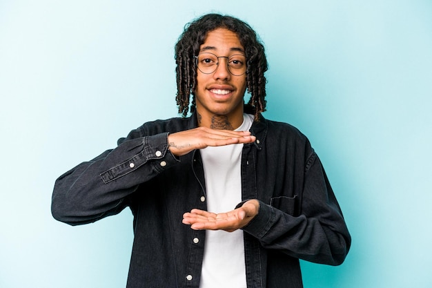 Young African American man isolated on blue background holding something with both hands product presentation