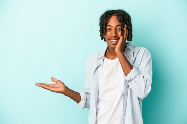 Young african american man isolated on blue background holds copy space on a palm, keep hand over cheek. Amazed and delighted.