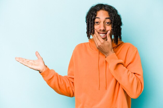 Young African American man isolated on blue background holds copy space on a palm keep hand over cheek Amazed and delighted