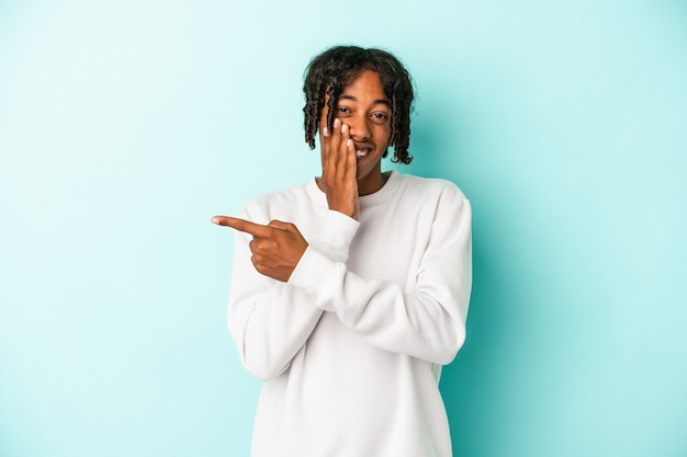 Young african american man isolated on blue background saying a gossip, pointing to side reporting something.