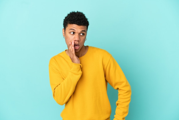 Young African American man isolated on blue background whispering something with surprise gesture while looking to the side