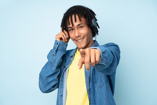 Young african american man isolated on blue wall listening music and pointing to the front