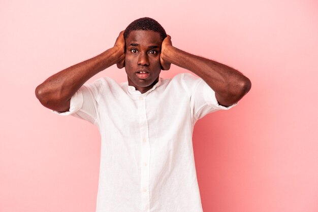 Young African American man isolated on pink background covering ears with hands trying not to hear too loud sound.