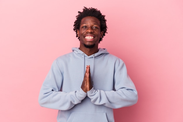 Young african american man isolated on pink background holding hands in pray near mouth, feels confident.