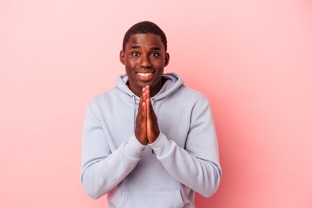 Young African American man isolated on pink background holding hands in pray near mouth, feels confident.