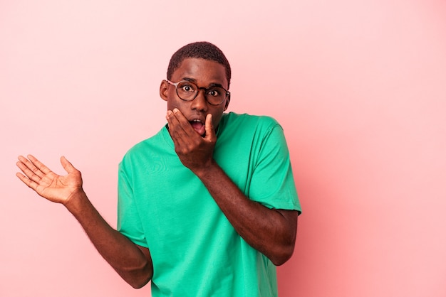 Young African American man isolated on pink background holds copy space on a palm, keep hand over cheek. Amazed and delighted.