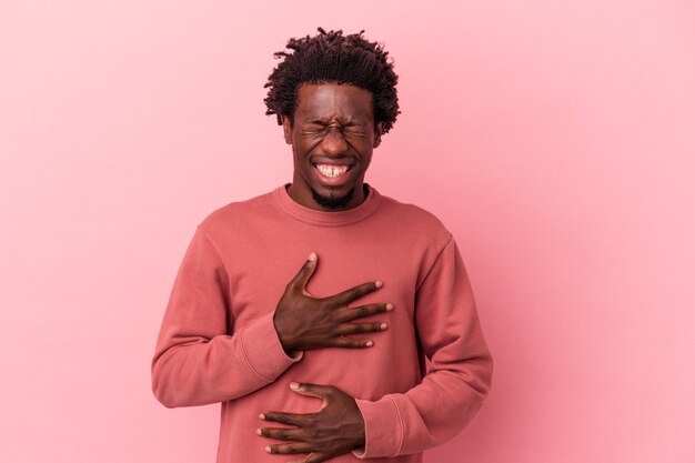 ピンクの背景に分離された若いアフリカ系アメリカ人の男は幸せに笑い、胃に手を保つことを楽しんでいます。