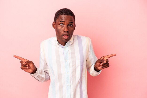 Young African American man isolated on pink background pointing to different copy spaces, choosing one of them, showing with finger.