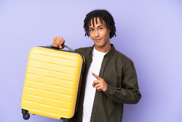 Young african american man isolated on purple background in vacation with travel suitcase