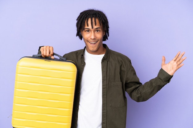 Young african american man isolated on purple in vacation with travel suitcase and surprised