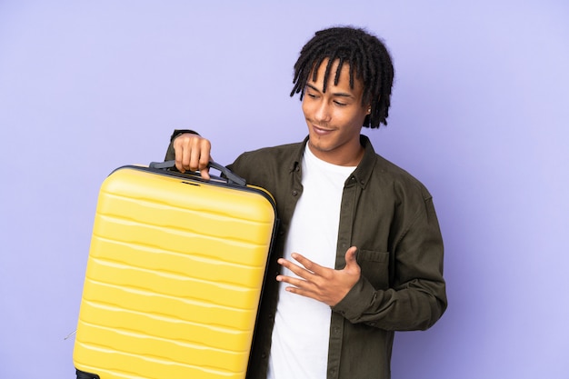 Young african american man isolated on purple wall in vacation with travel suitcase