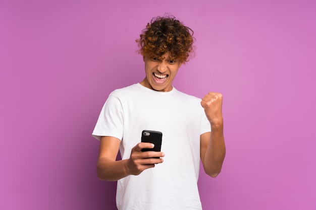 Giovane uomo afroamericano sopra isolato muro viola con il telefono in posizione di vittoria