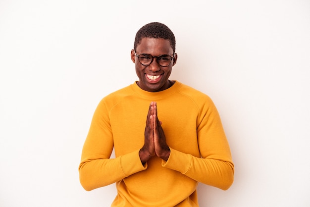 Young African American man isolated on white background holding hands in pray near mouth, feels confident.