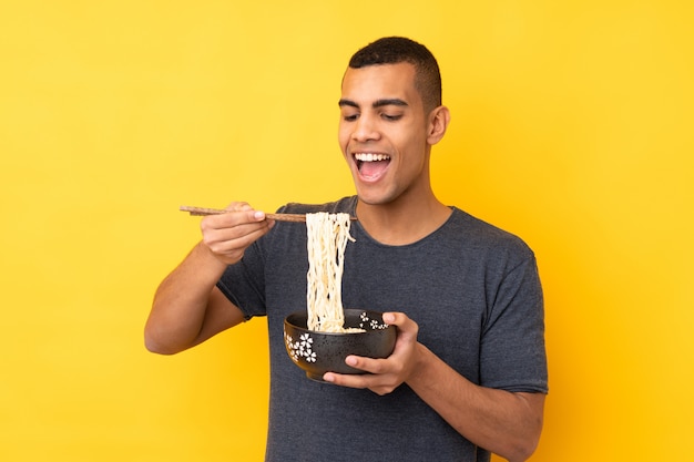 Young African American man over isolated yellow wall holding a bowl of noodles with chopsticks and eating it