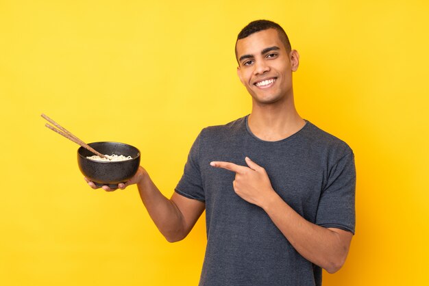 Giovane uomo afroamericano sopra il muro giallo isolato e puntandolo mentre si tiene una ciotola di noodles con le bacchette