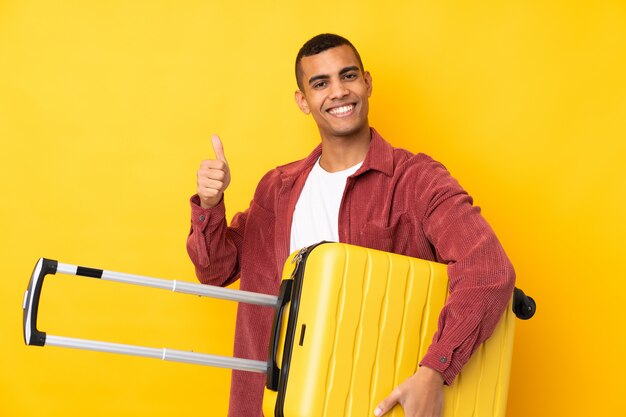 Young African American man over isolated yellow wall in vacation with travel suitcase and with thumb up