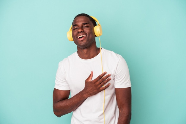 Young African American man listening to music isolated on blue background laughs out loudly keeping hand on chest.