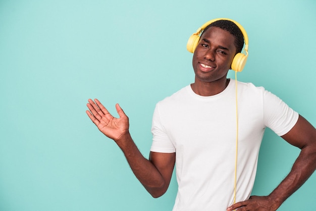 Giovane uomo afroamericano che ascolta musica isolata su sfondo blu che mostra uno spazio di copia su un palmo e tiene un'altra mano sulla vita.