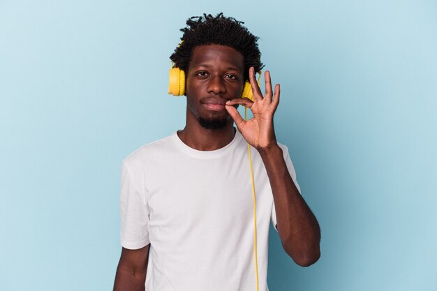 Young african american man listening to music isolated on blue background with fingers on lips keeping a secret.