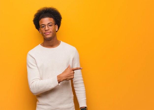 Young african american man over an orange wall smiling and pointing to the side