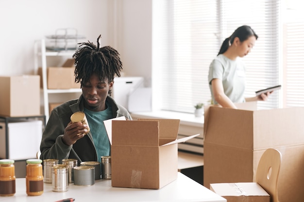 Young African-American man packing canned food to boxes at charity and donations event, copy space