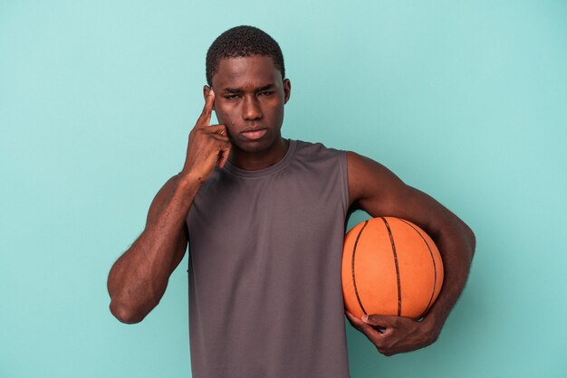 Young African American man playing basketball isolated on blue background pointing temple with finger, thinking, focused on a task.