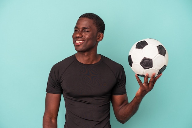 Young African American man playing football isolated on blue background looks aside smiling, cheerful and pleasant.