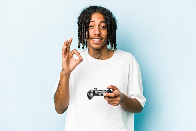 Young African American man playing with a video game controller isolated on blue background cheerful and confident showing ok gesture