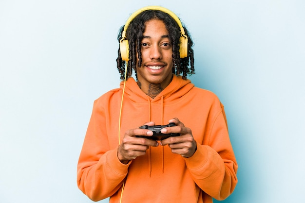 Young African American man playing with a video game controller isolated on blue background happy smiling and cheerful