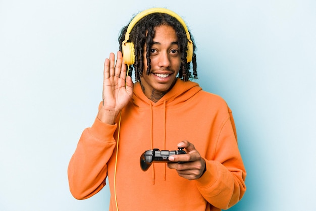 Young African American man playing with a video game controller isolated on blue background trying to listening a gossip