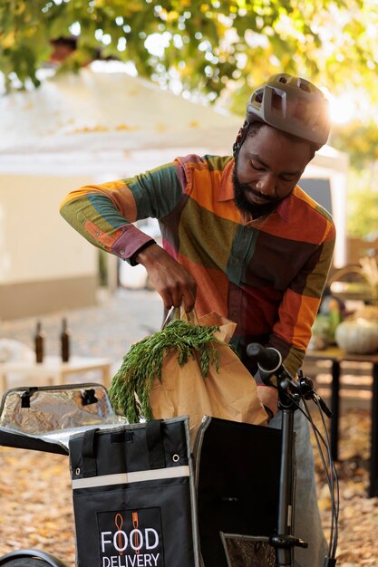 Молодой афроамериканец кладет биоорганические продукты в термосумку, забирает заказы с фермерского рынка и катается на велосипеде. Доставка овощей и фруктов местного производства клиентам с фермы.