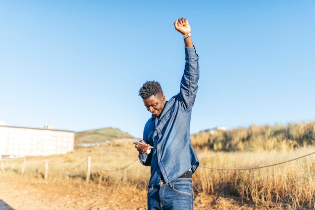 Young african american man raising fist in victory sign looking at smartphone on sunny day