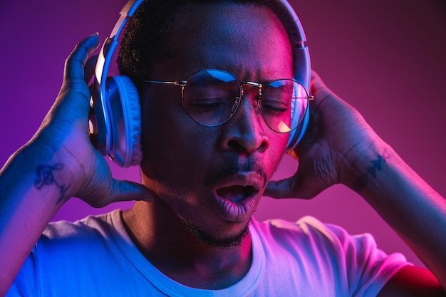 Young african-american man's listening to music in headphones in neon light on gradient background. Male portrait. Concept of human emotions, facial expression, summer holidays or weekend, hobby,